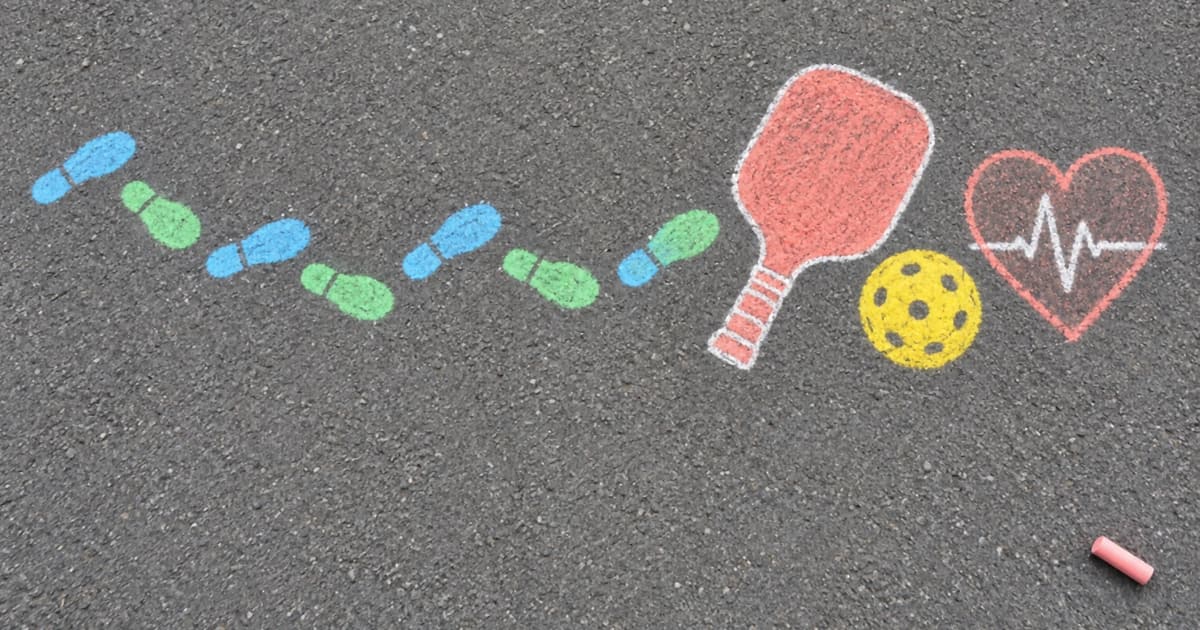 Sidewalk chalk drawing on asphalt of a pickleball paddle, a ball, footprints and a heart with a heartbeat line